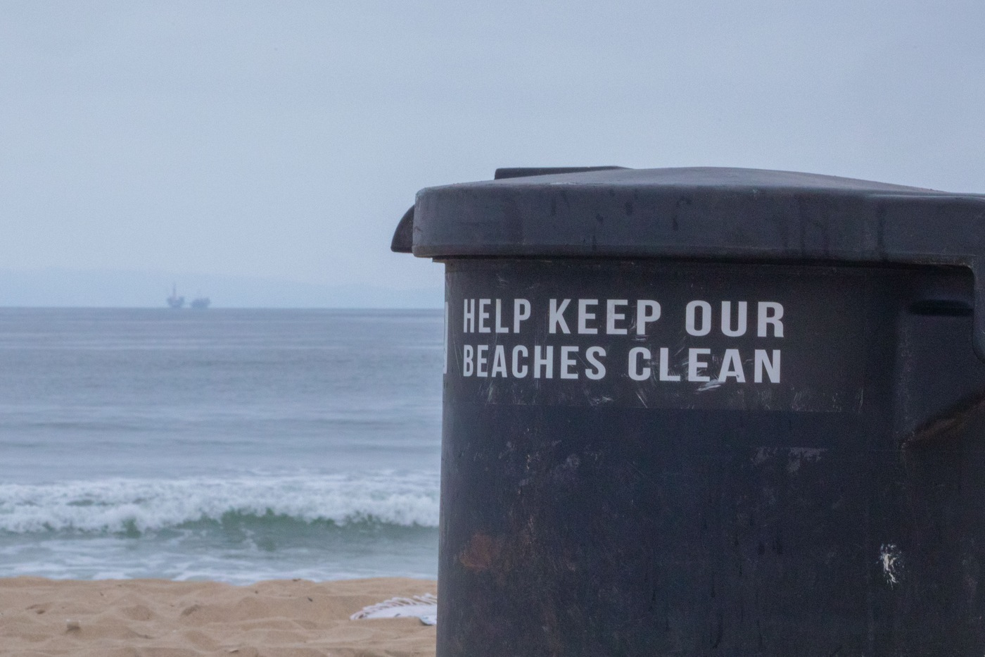 A waste disposal container at Huntington Beach with "Keep Our Beaches Clean" posted on it. In the distance, an oil rig can be seen.
