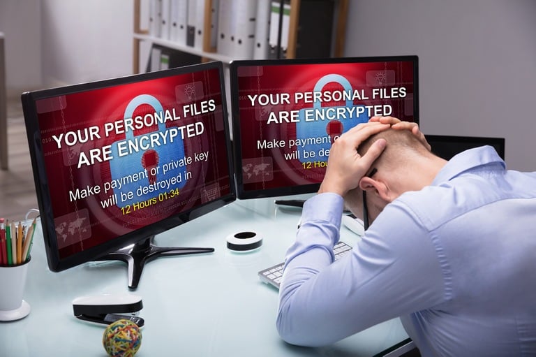 Stock image depicting distressed man in front of computer monitors displaying ransomware lock screens.