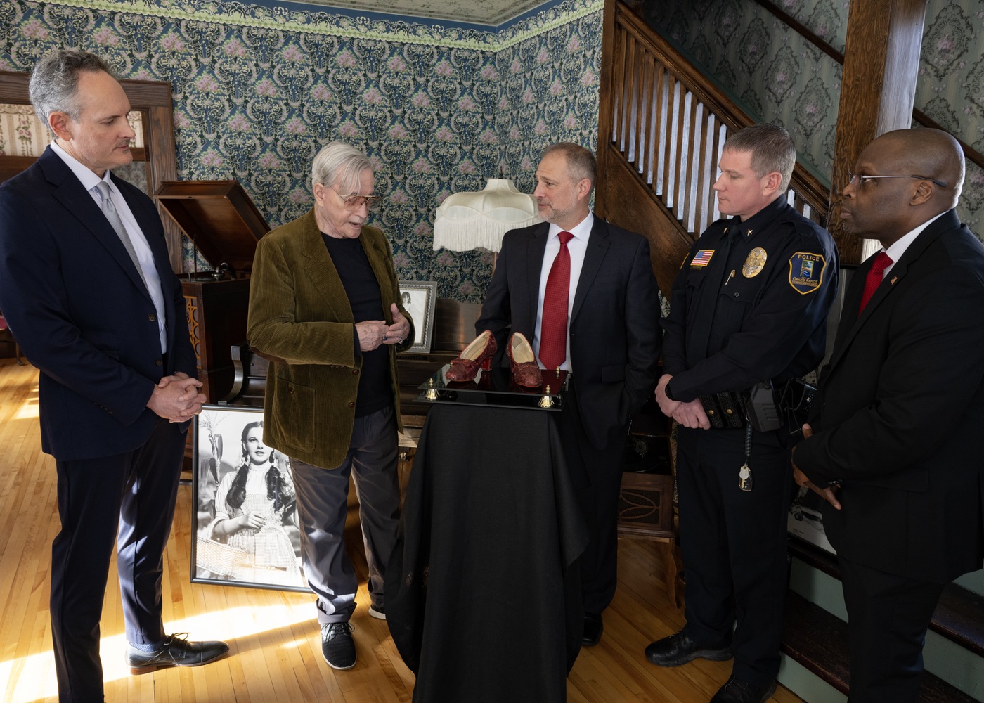 This photo shows (from left to right) Brian Chanes from Heritage Auctions, ruby slippers owner Michael Shaw, Special Agent Christopher Dudley of FBI Minneapolis, Grand Rapids Police Department Chief Andy Morgan, and FBI Minneapolis Special Agent in Charge Alvin M. Winston, Sr., as Shaw is joyfully reunited with the ruby slippers after nearly a decade. The event unfolded in Judy Garland’s childhood home at the Judy Garland Museum in Grand Rapids, Minnesota. Placed atop the very pedestal from which they were stolen in 2005, the return of the slippers provided a poignant moment of closure.