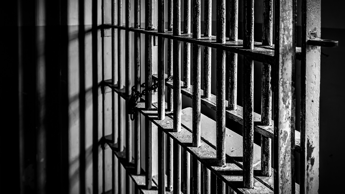 Black and white stock image depicting the barred doors of a prison cell with a chain and lock.